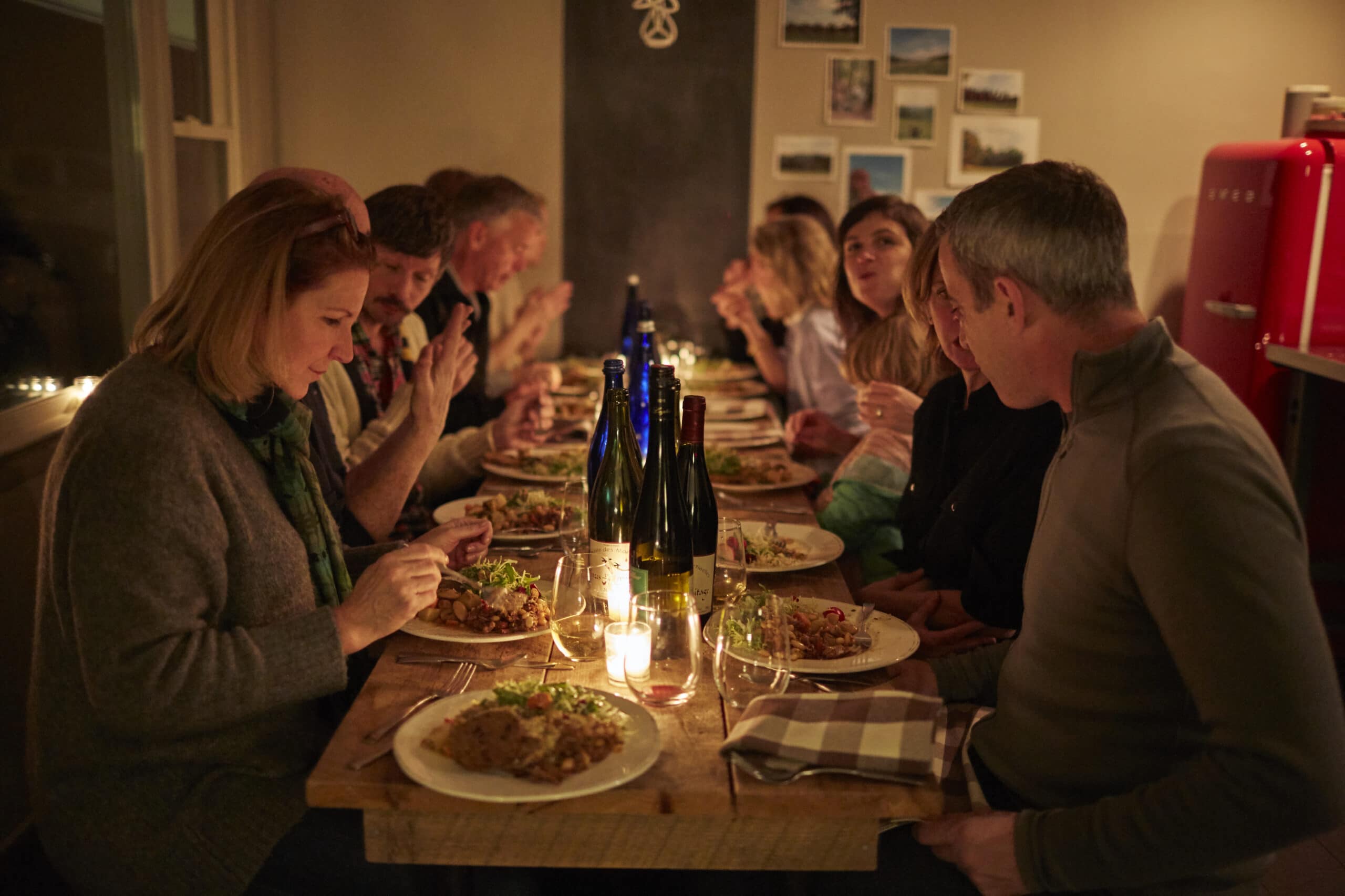 Group of adults enjoying a dinner party at a table with candlelight in a cozy, dimly lit motel in the Berkshires, sharing conversation and food.